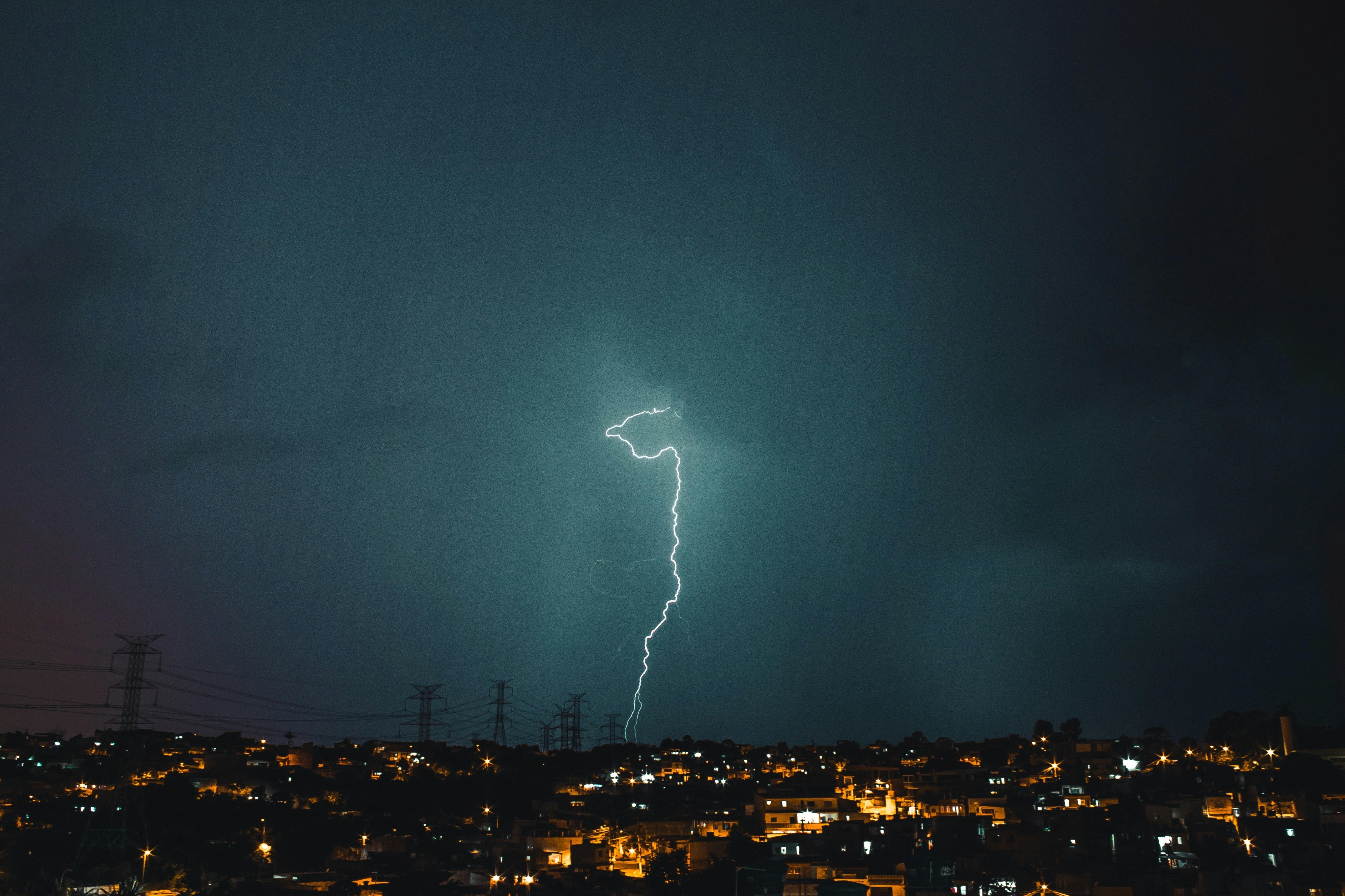 Striking lightning bolt amid dark storm clouds above city skyline, captured at night.