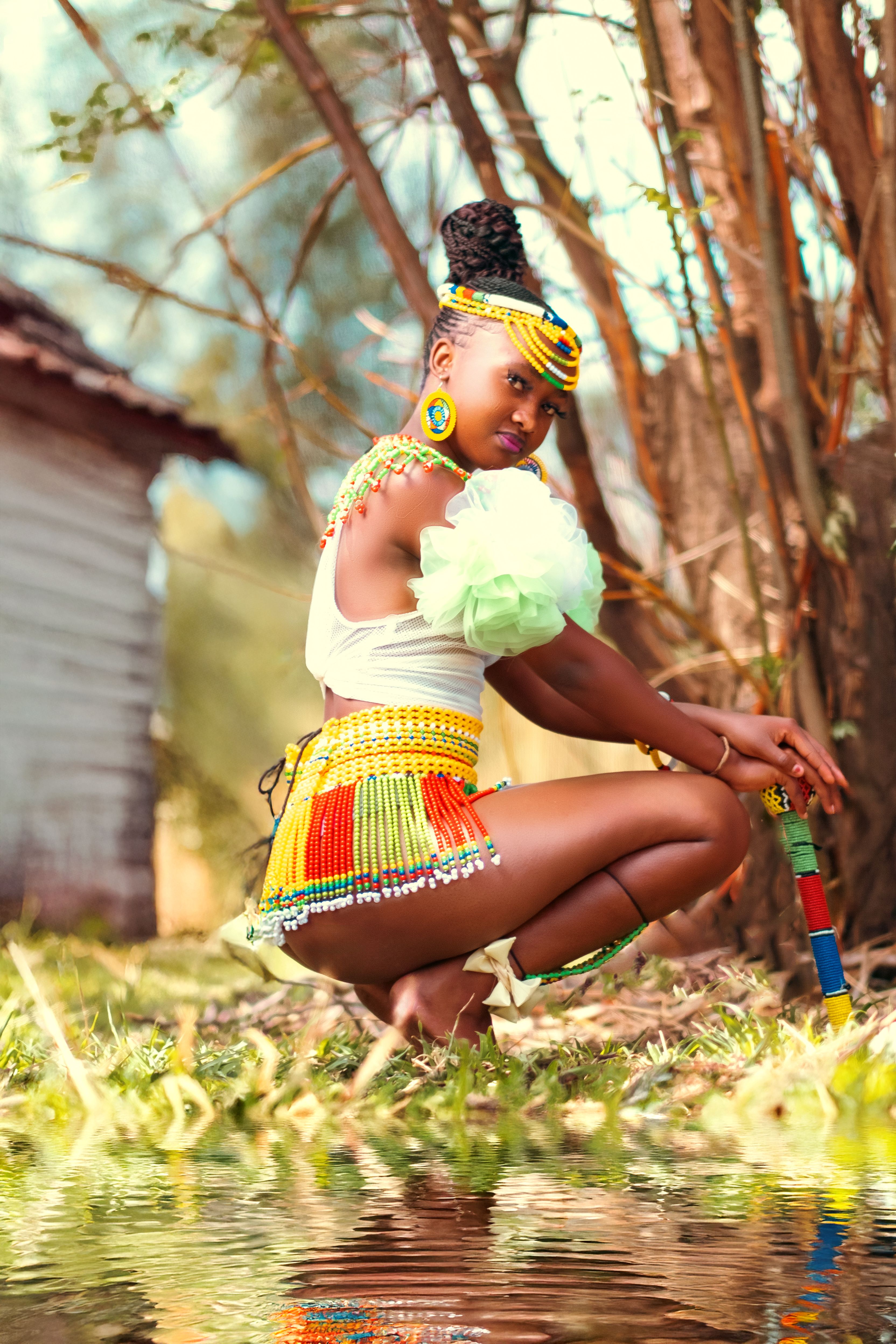 A woman in colorful African traditional attire kneels outdoors by a reflective water surface.