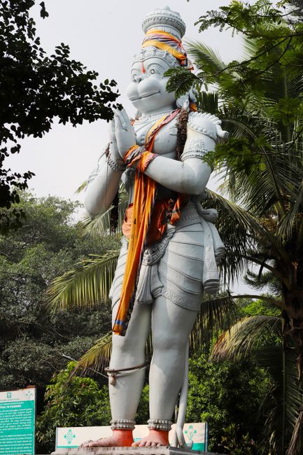 A large statue of Hanuman in prayer pose surrounded by tropical trees and foliage.