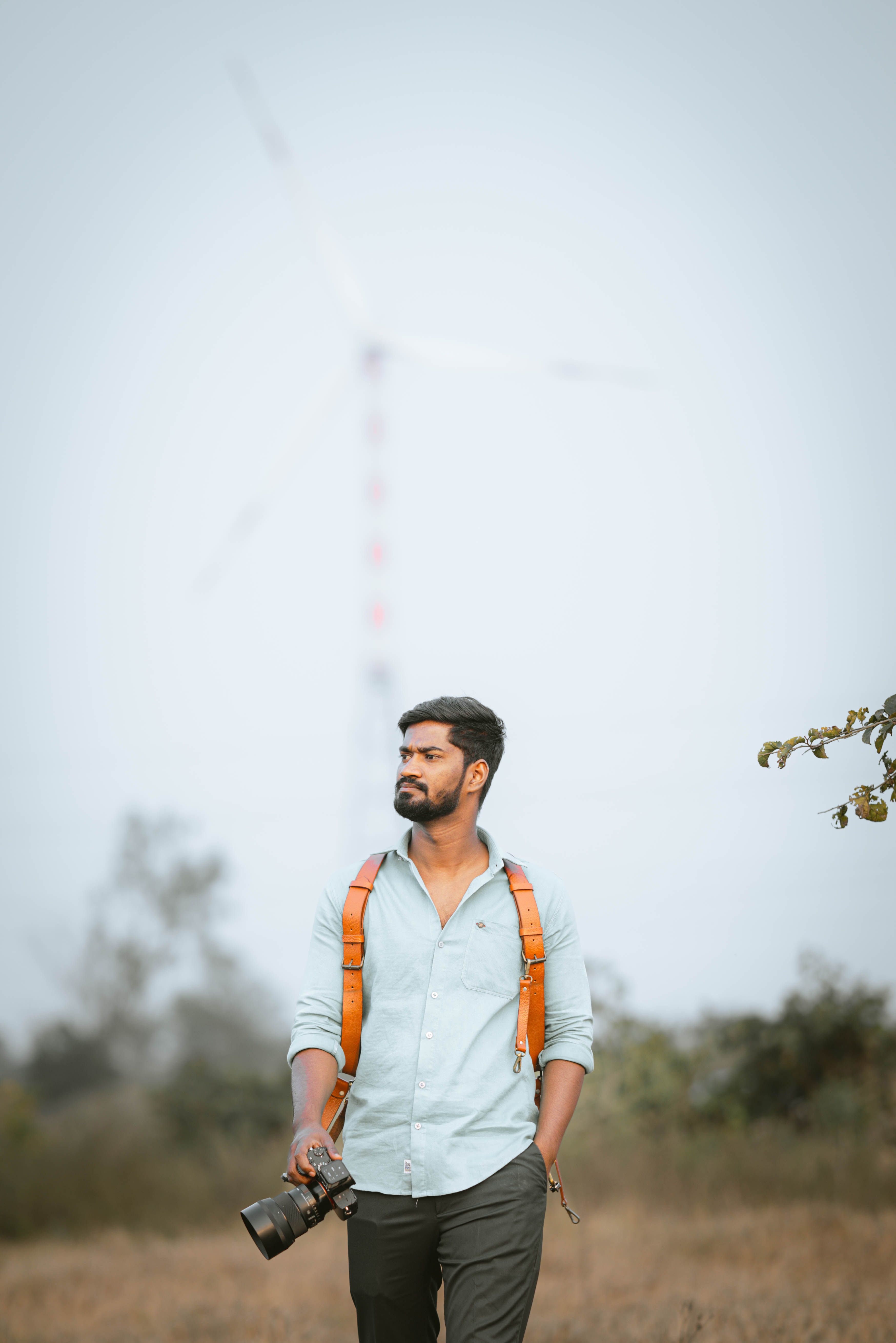 Man with a backpack holding a camera, walking outdoors near a wind turbine.