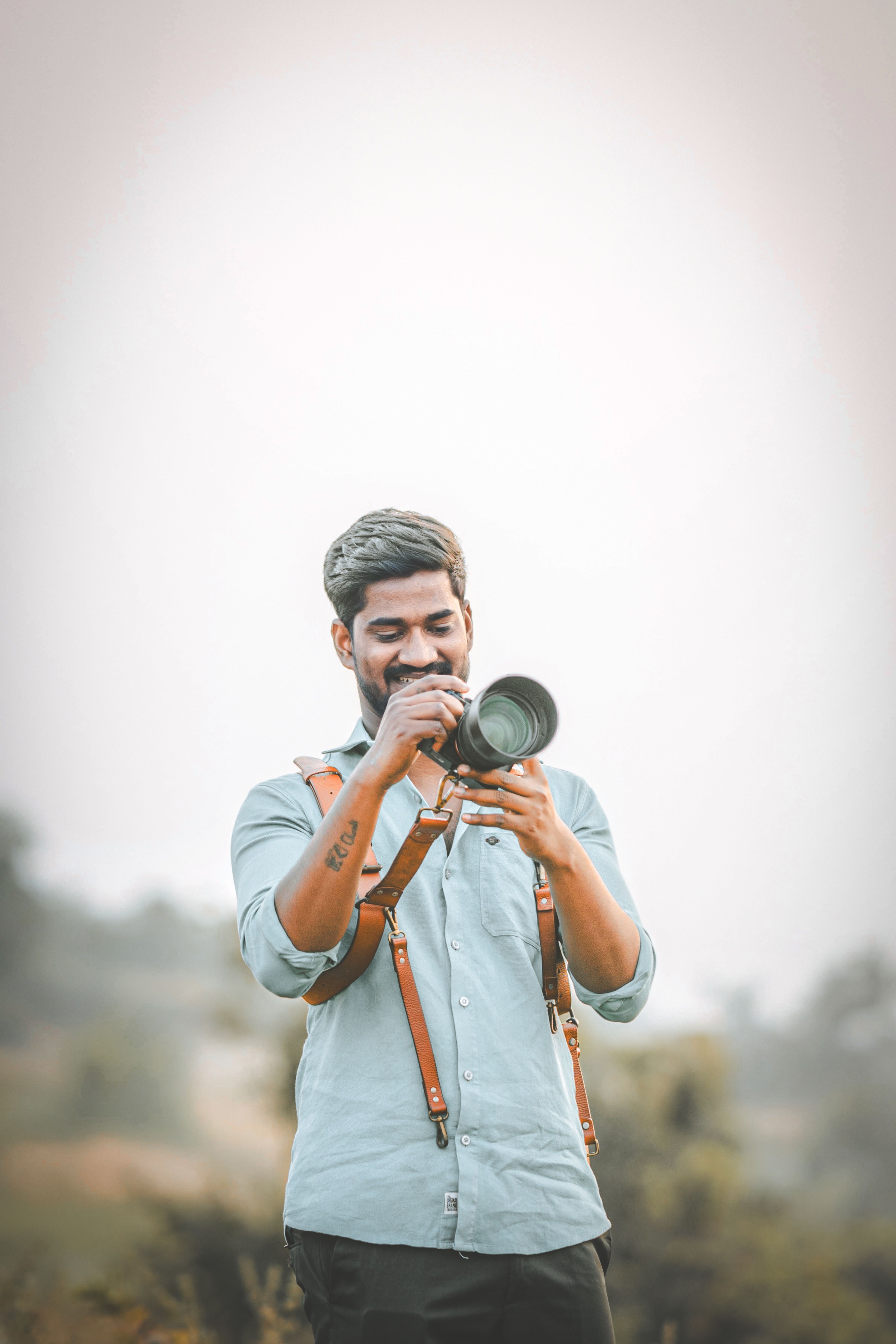 Man with a camera outdoors, focusing, wearing strap and blue shirt.