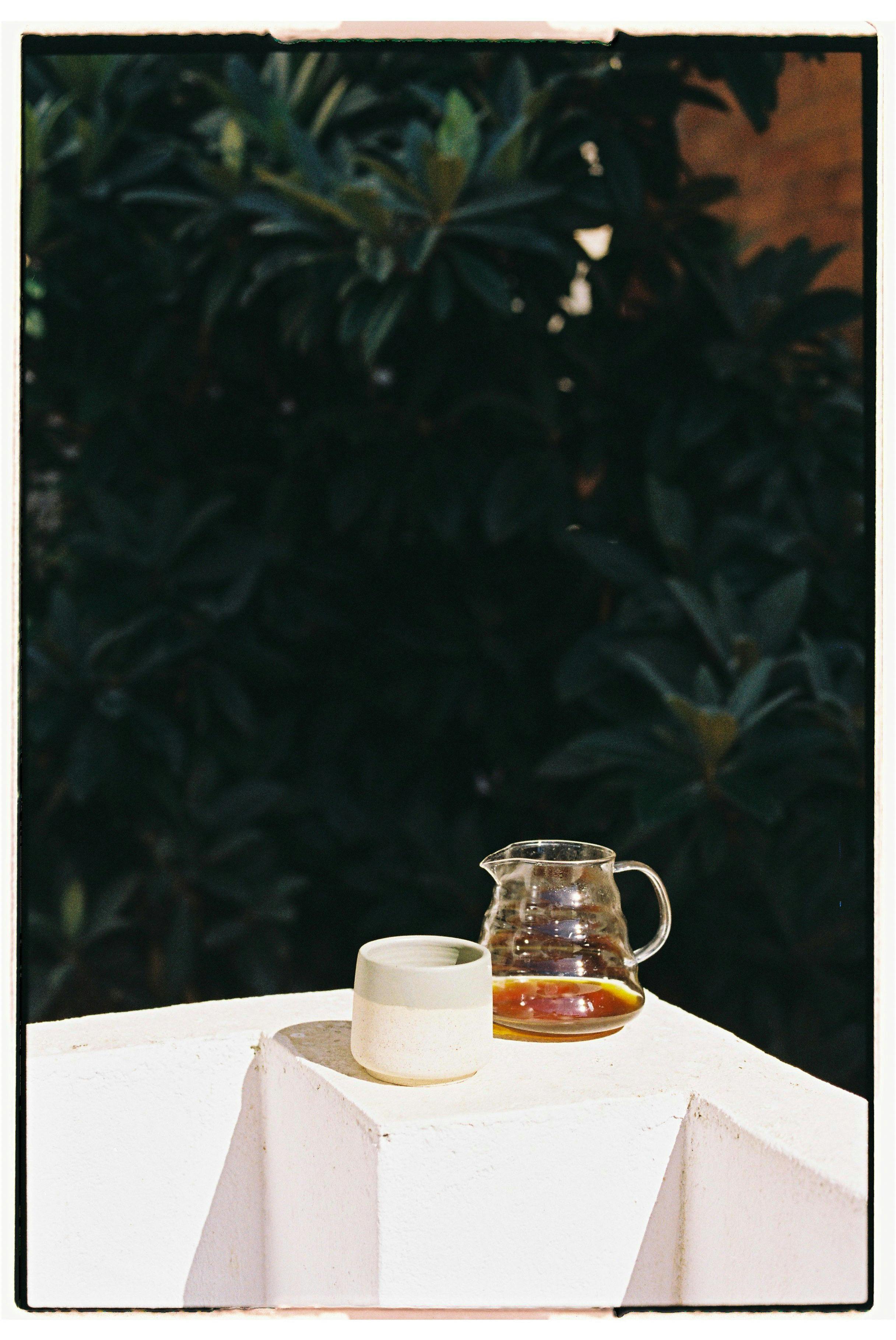 A glass carafe and ceramic cup on a white surface with sunlight outdoors.