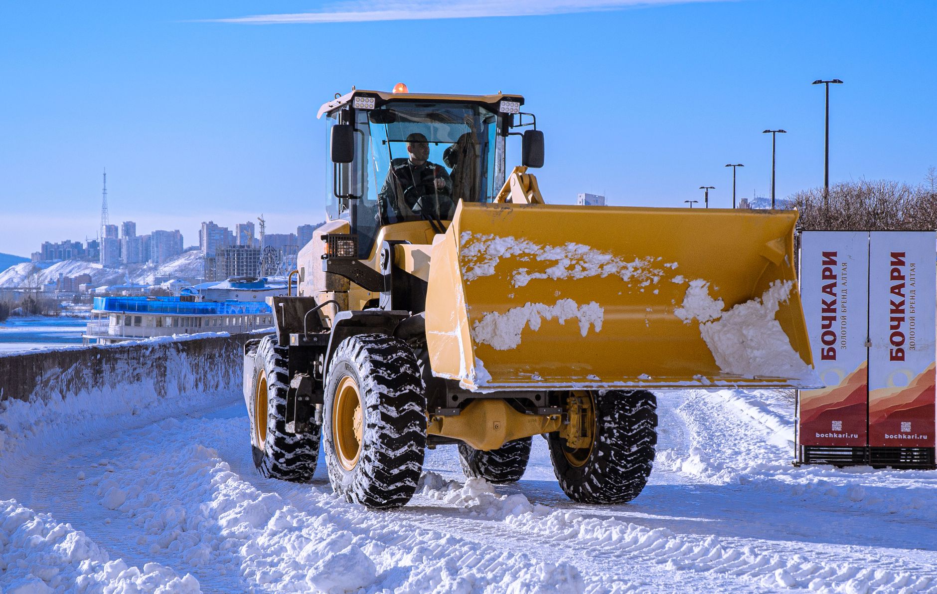 Yellow snowplow clearing snow on urban road with city skyline in background.