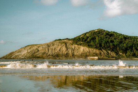 A peaceful coastal scene featuring waves and a rocky hill under a clear sky.