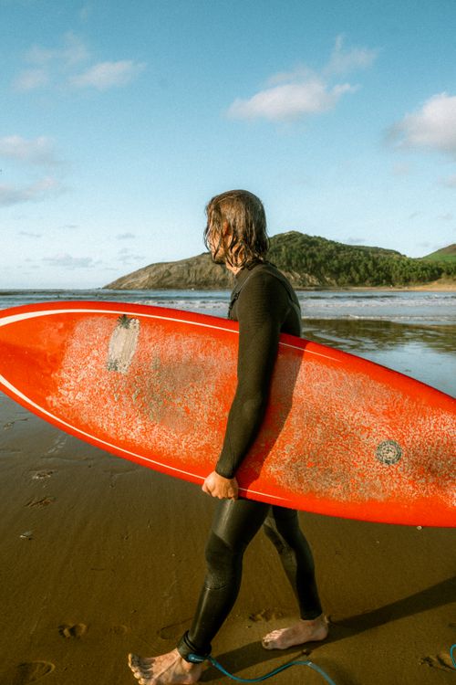 Free Adult surfer carrying a red board along a sunny beach, embracing the ocean waves. Stock Photo