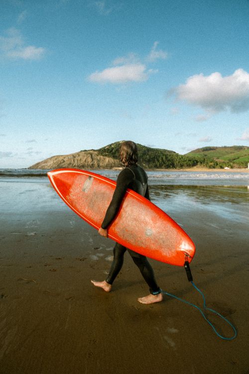 Free Male surfer with red surfboard walking along a scenic beach, ready to catch waves on a sunny day. Stock Photo