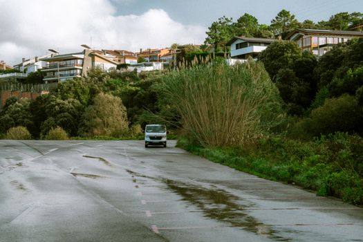 A serene suburban scene featuring a parked van on a wet road surrounded by lush greenery and modern houses.