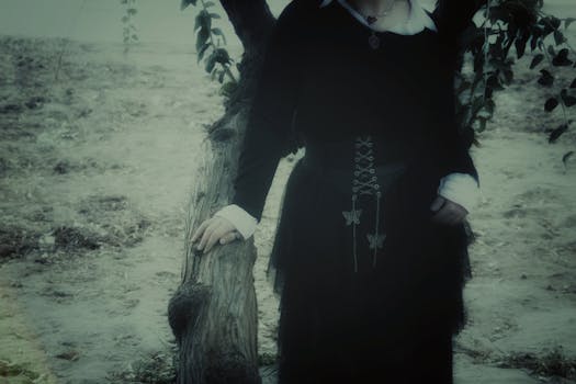 Gothic woman in dark attire leaning against tree, mystical ambiance outdoors.