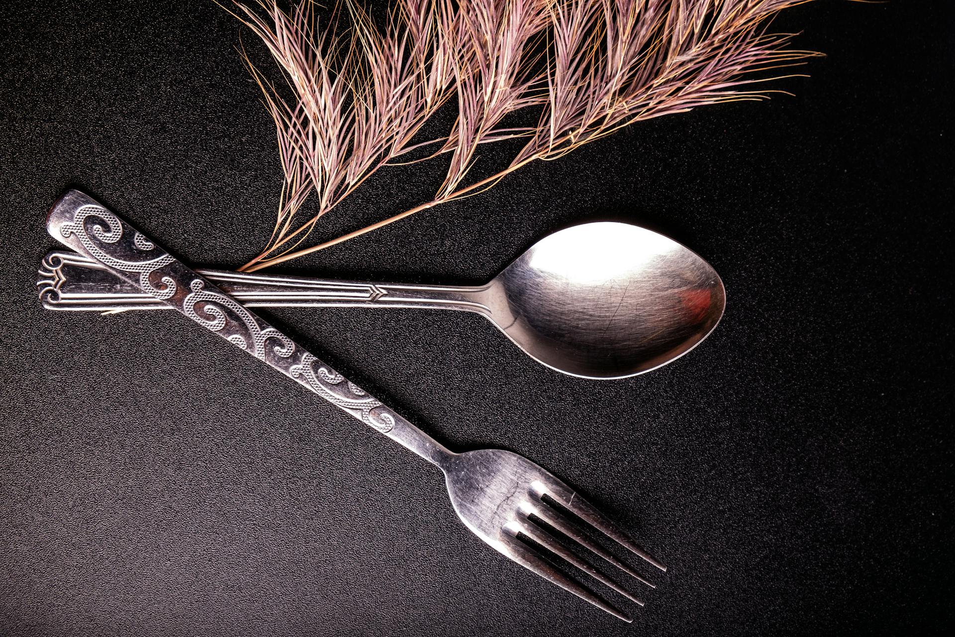 Flat lay of elegant silver fork and spoon with decorative design beside dried ornamental grass.