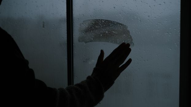 Silhouette of a hand touching a wet, misted window during rain, creating an emotional ambiance.