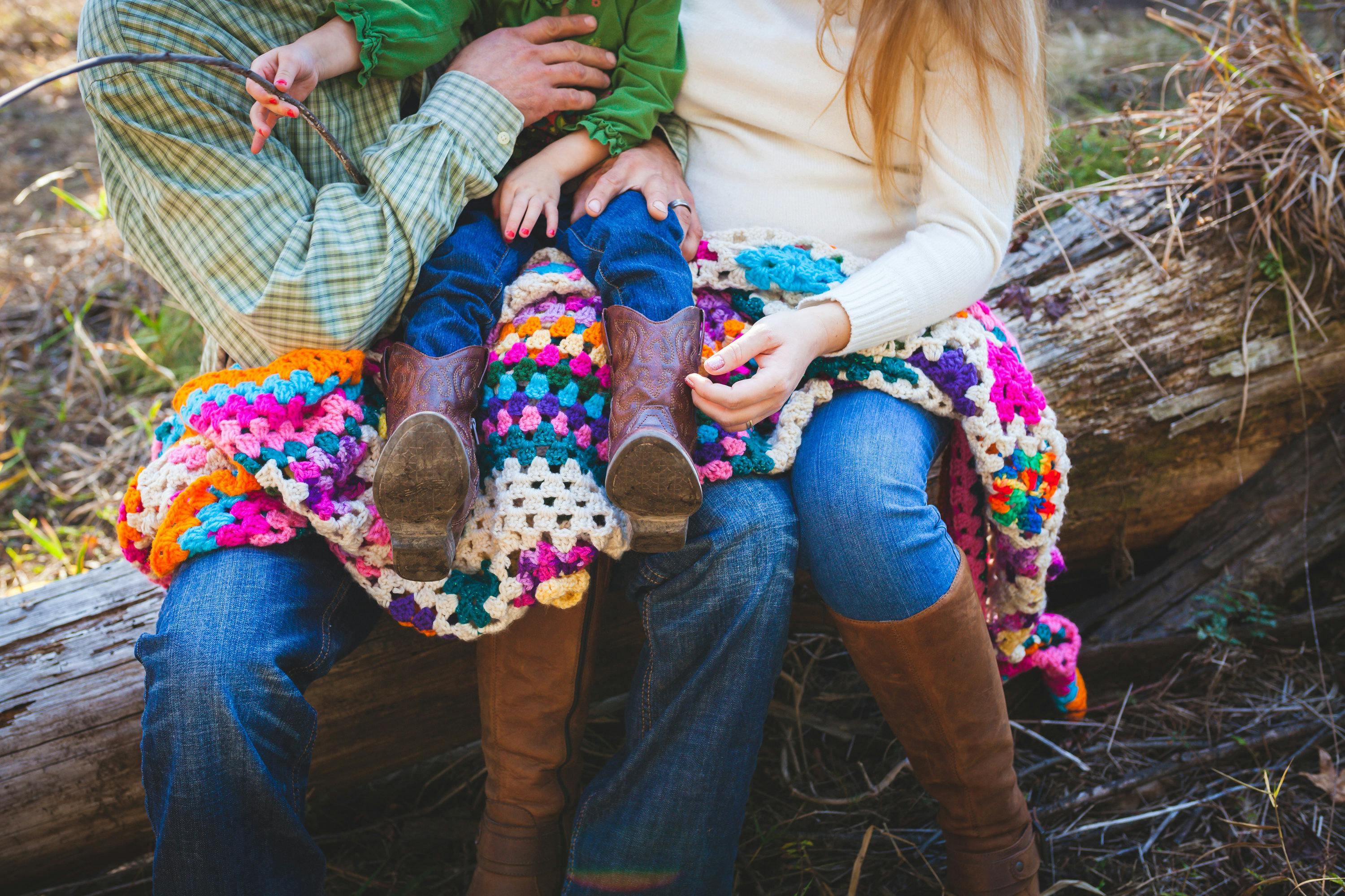 A family enjoys a peaceful day outdoors sitting on a log with a colorful crochet blanket.