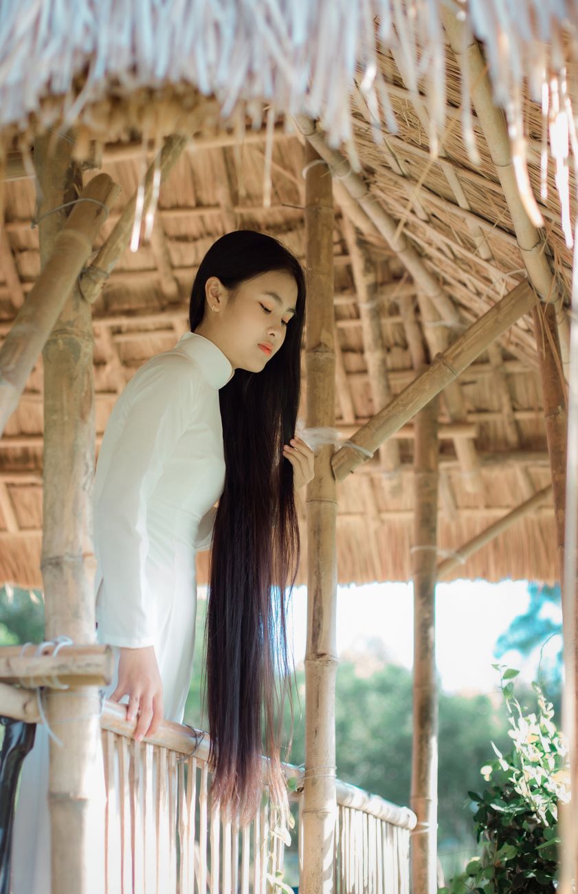 Vietnamese woman in white áo dài under bamboo structure in sunlight.