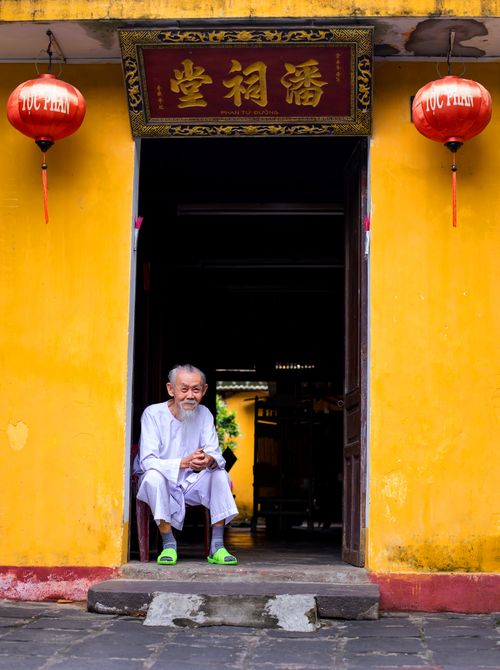 Free Elderly man sitting at a vibrant yellow temple entrance in Hoi An, Vietnam. Stock Photo
