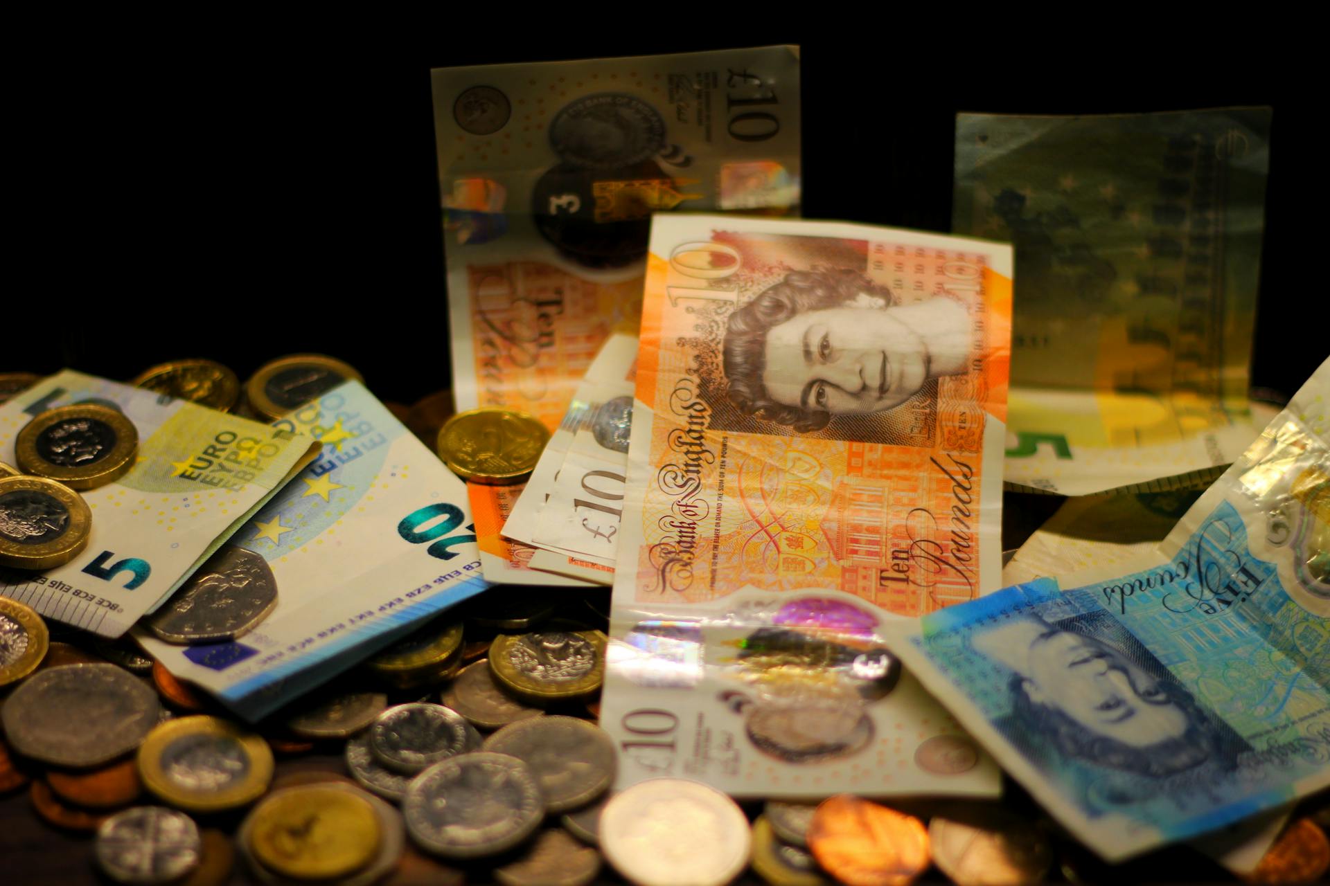 A dark-toned close-up of various Euro and Pound banknotes and coins scattered.