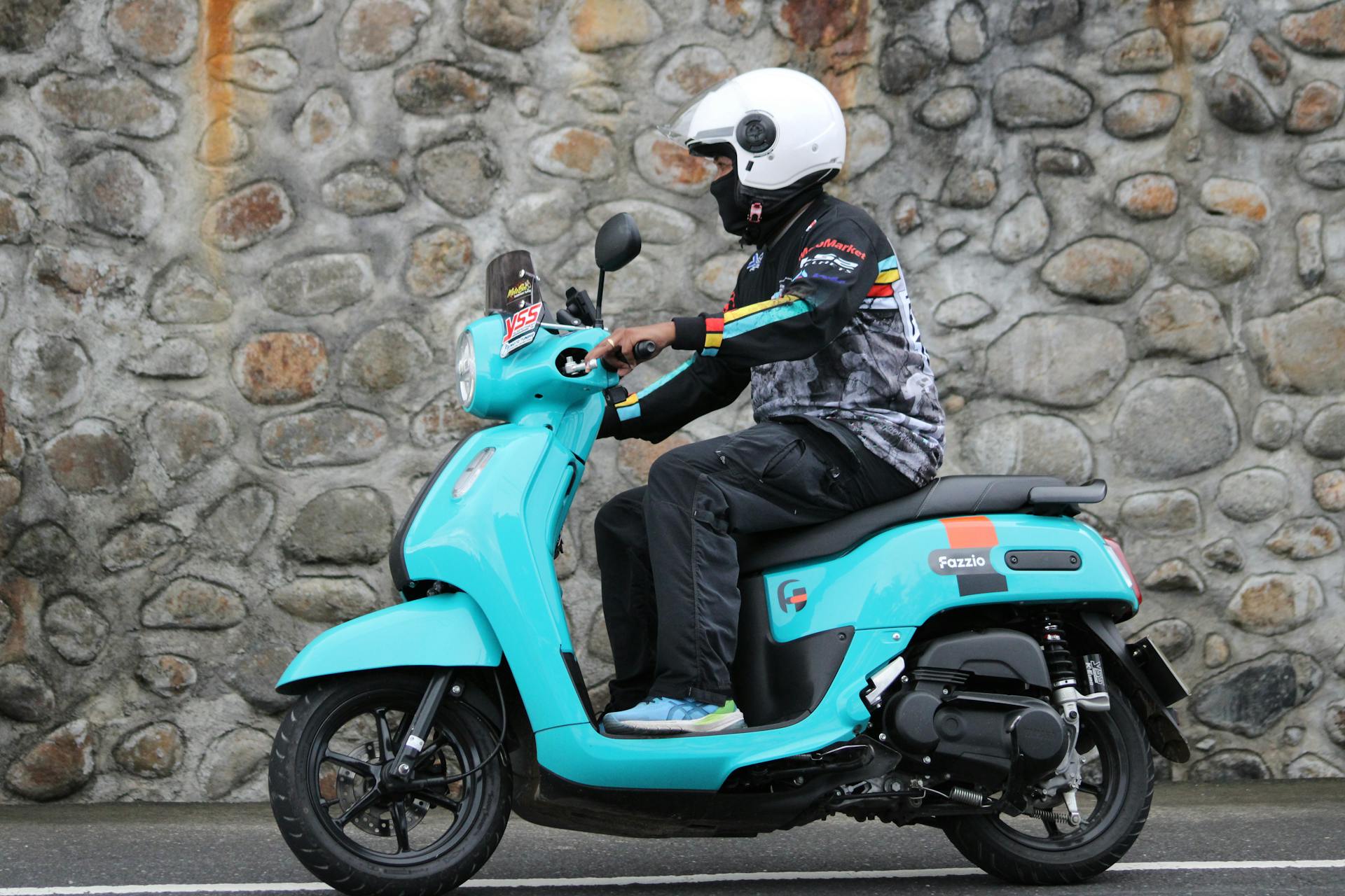 Individual wearing helmet on teal scooter near a stone wall, street view.