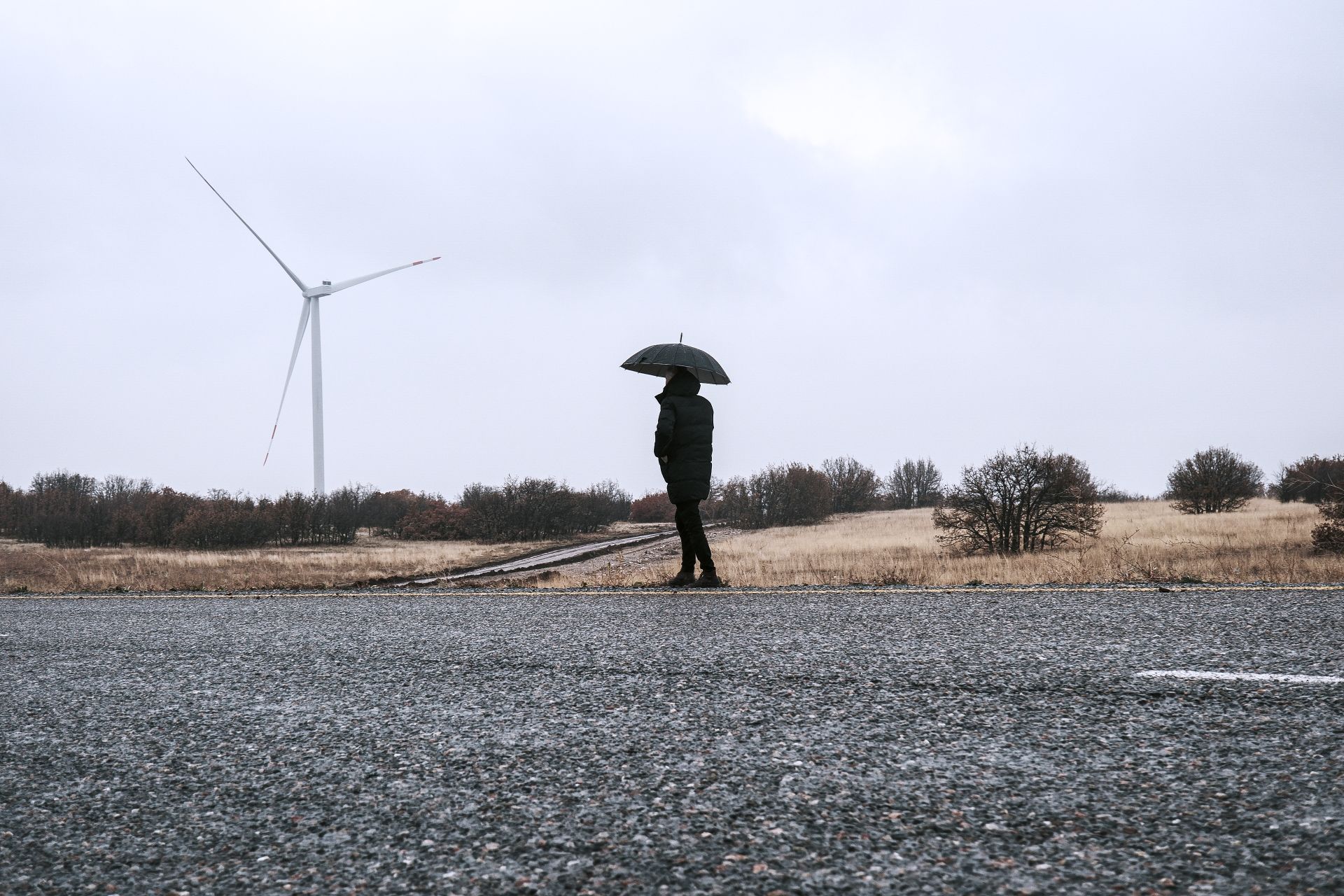 A person with an umbrella walks on a rainy day beside a wind turbine in the countryside.