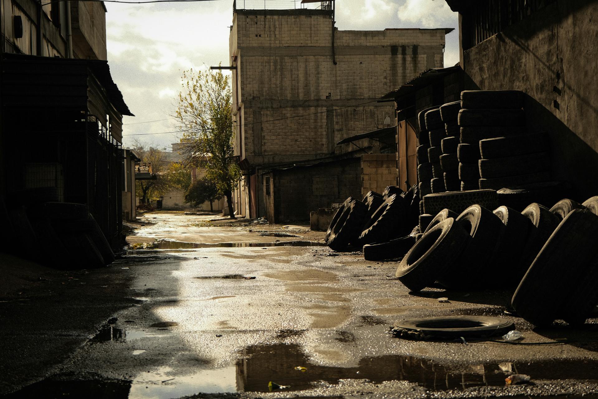 A deserted urban alley lined with stacked tires under a dramatic sunset sky, reflecting on the wet ground.