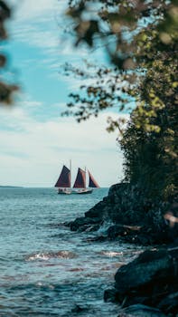 A serene seascape featuring a sailboat with red sails navigating along a rocky coastline.