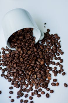 A white mug tipped over, spilling roasted coffee beans on a white surface.
