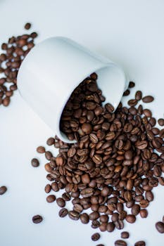 Brown coffee beans spilling from a white cup onto a white surface, showcasing richness and aroma.