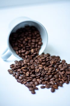 Close-up of spilled coffee beans from a white mug onto a white surface, creating a dynamic scene.