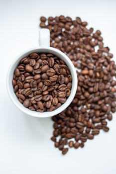 High-angle view of a white cup filled with coffee beans on a white surface.