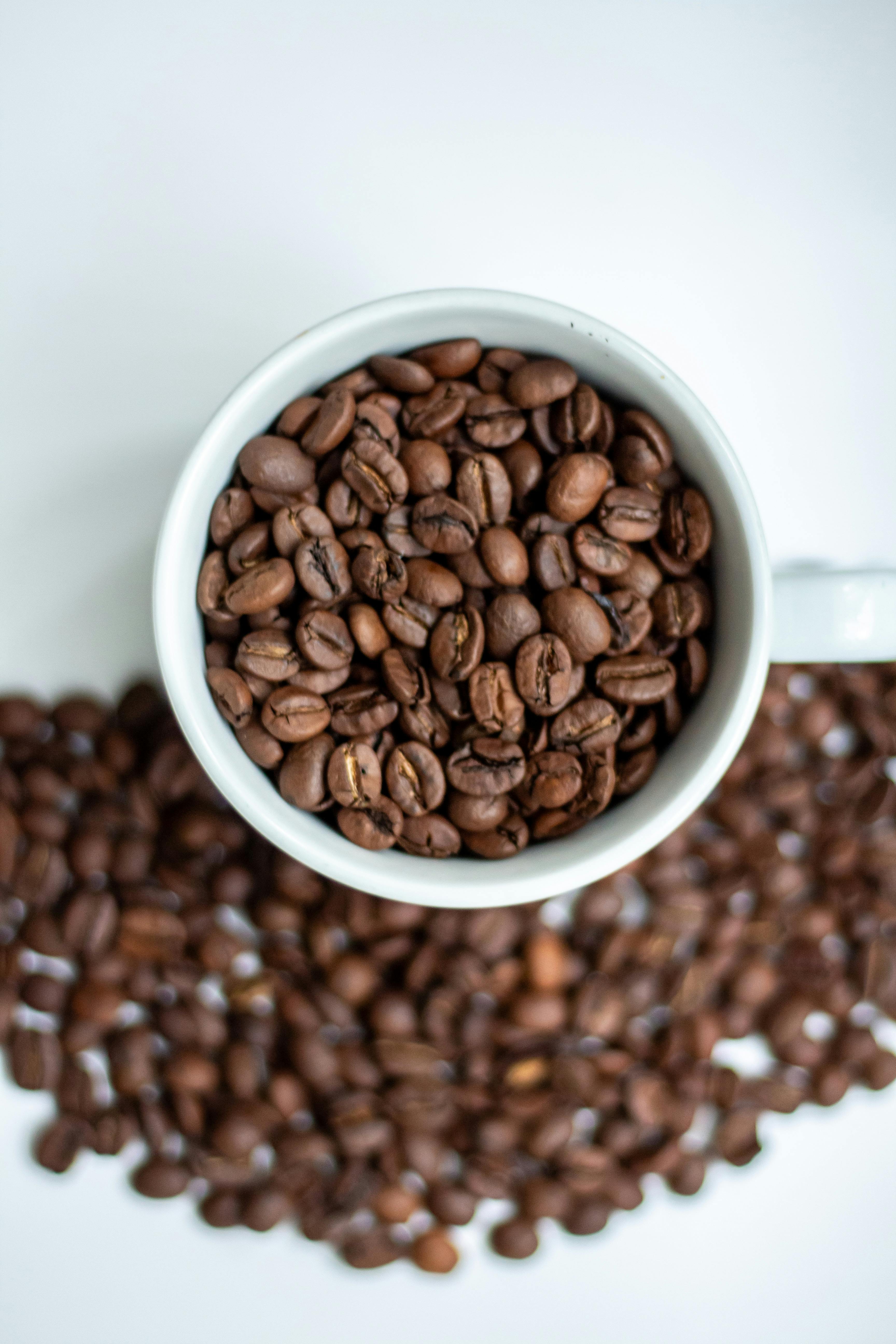 Top view of coffee beans in a cup with some beans spilled on a white surface.