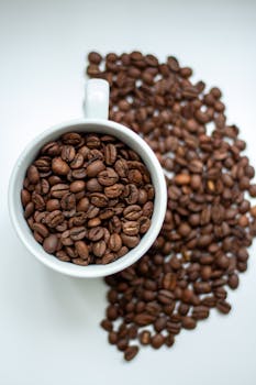 Aesthetic top view of coffee beans filling and overflowing from a white mug on a white background.