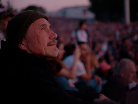 An elderly man sits among a crowd, captivated by an outdoor concert at twilight in Bratislava. - Fotografer Sidoarjo