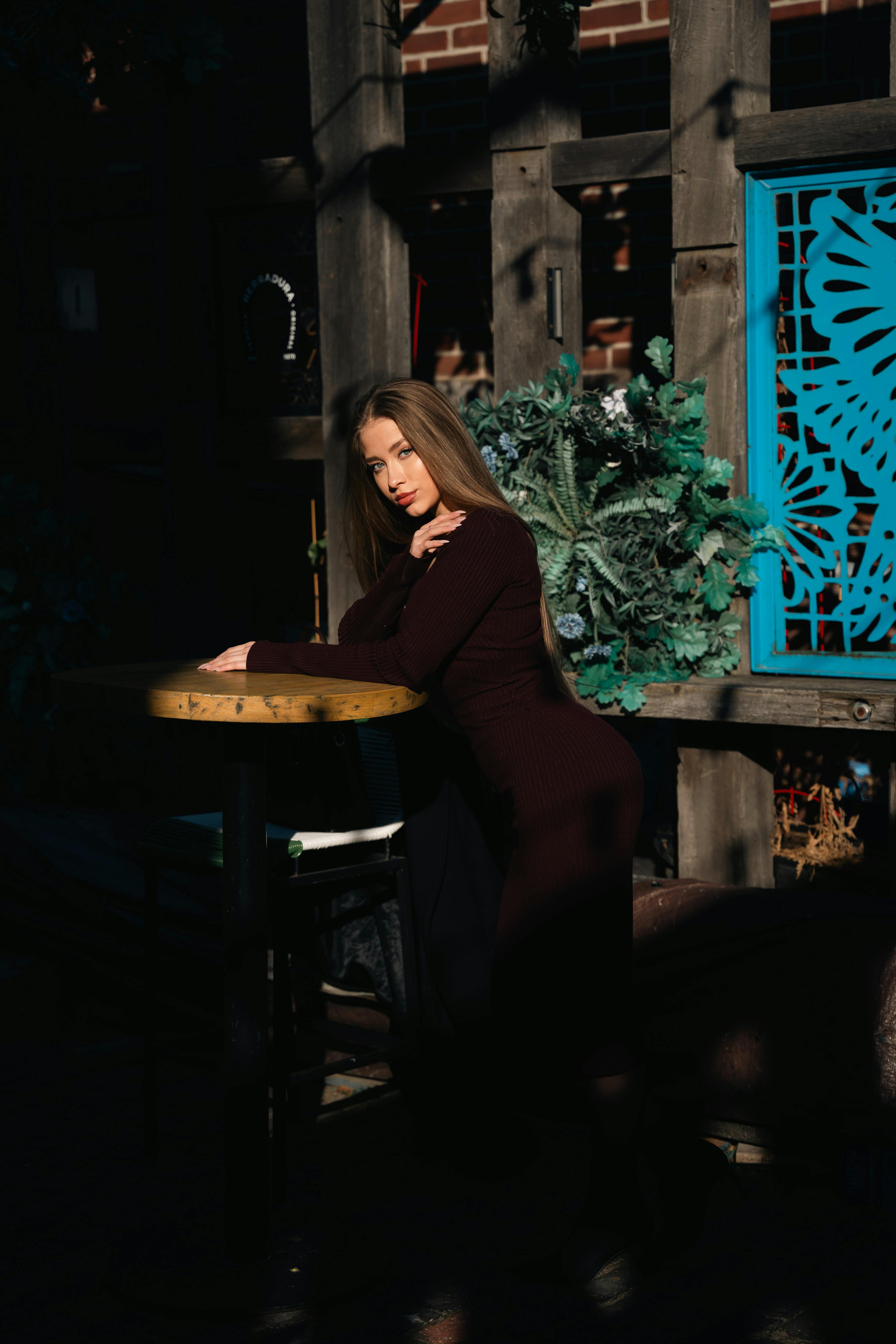 gratis Een vrouw in een donkere jurk poseert peinzend bij een tafeltje op het terras in een stedelijke omgeving, met een artistieke blauwe achtergrond. Stockfoto