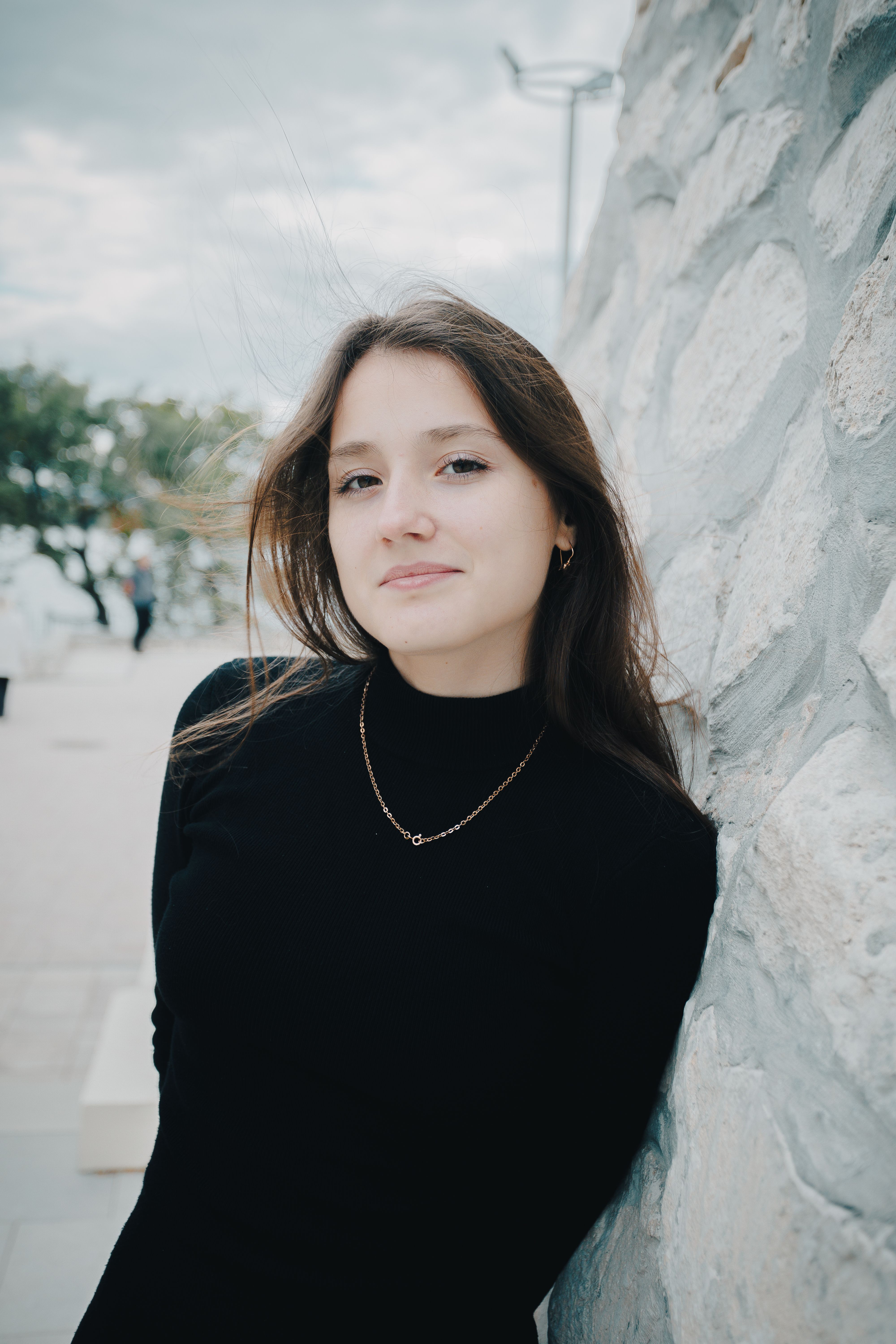 Young woman in black sweater leaning on a stone wall, smiling outdoors.