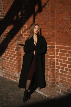 Fashionable woman in black coat stands confidently against a red brick wall in sunlight.