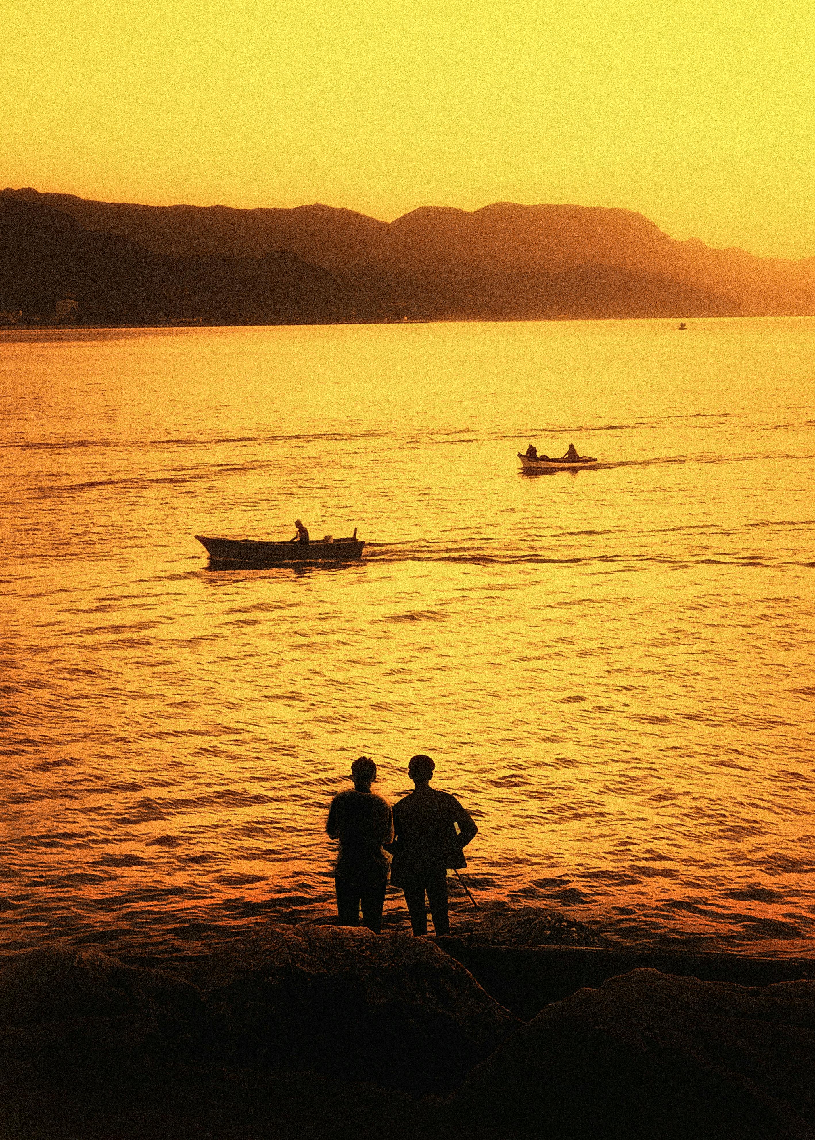 Two silhouettes admire a golden sunset over Cide Bay, Türkiye as boats glide by.