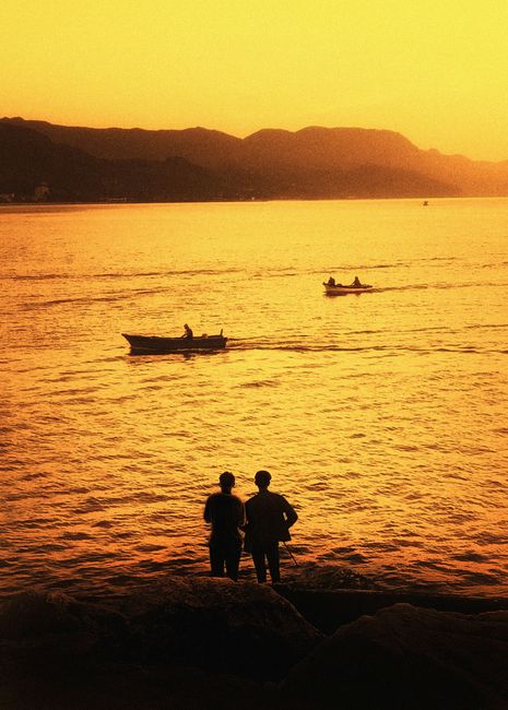 Two silhouettes admire a golden sunset over Cide Bay, Türkiye as boats glide by.