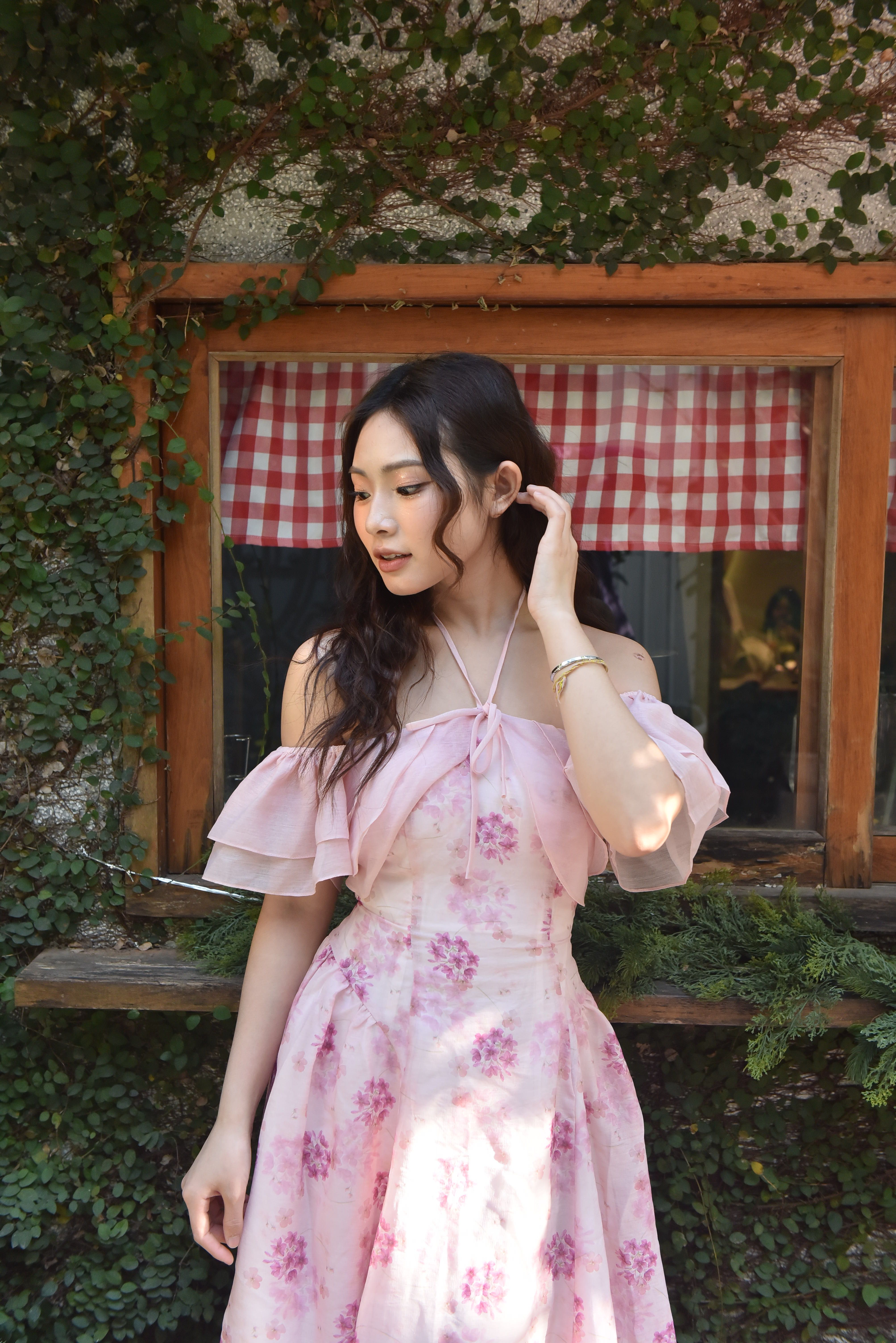 A young woman in a pink floral dress poses gracefully by a window with ivy.