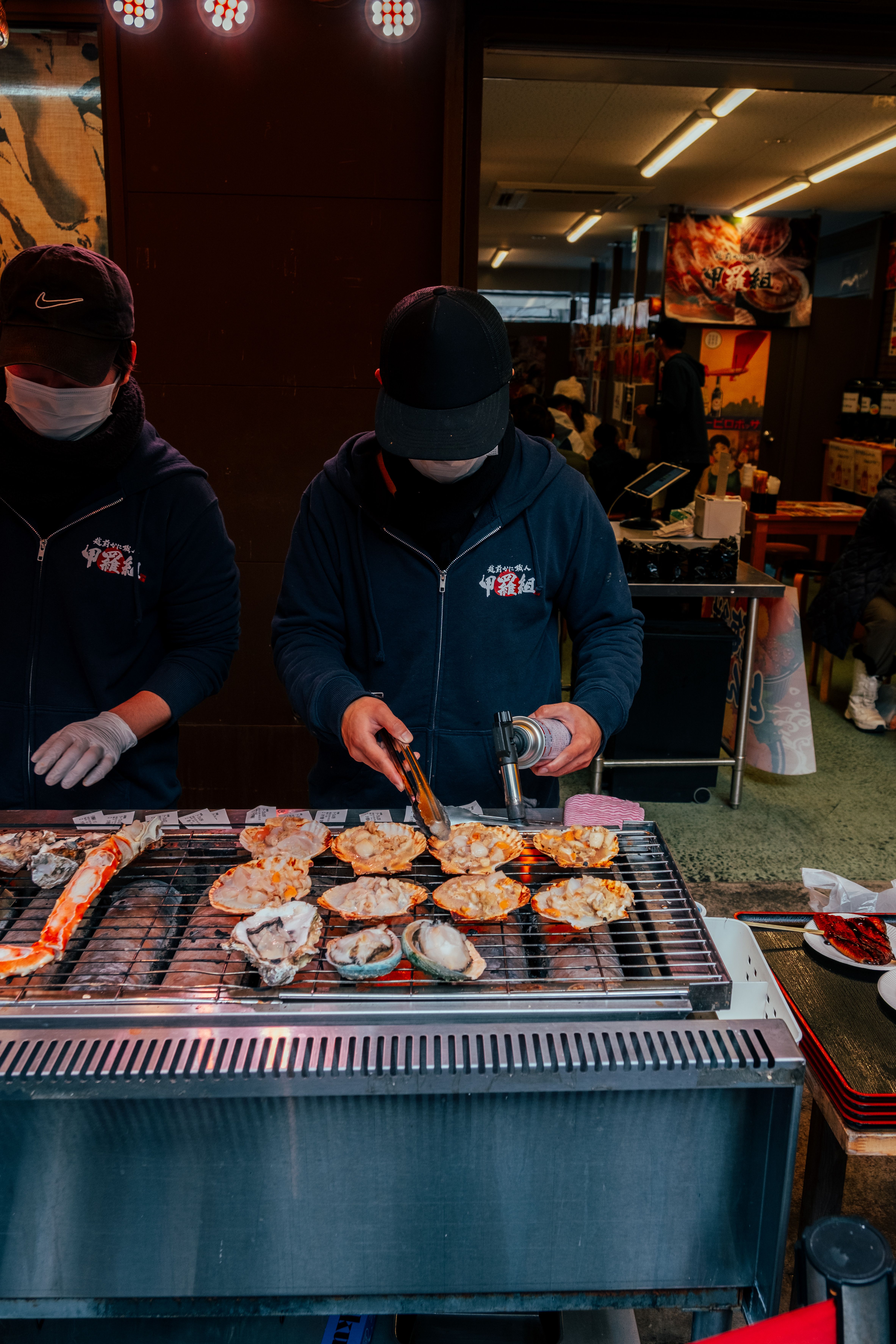Two vendors prepare grilled seafood at a bustling street food stall in Tokyo, Japan.