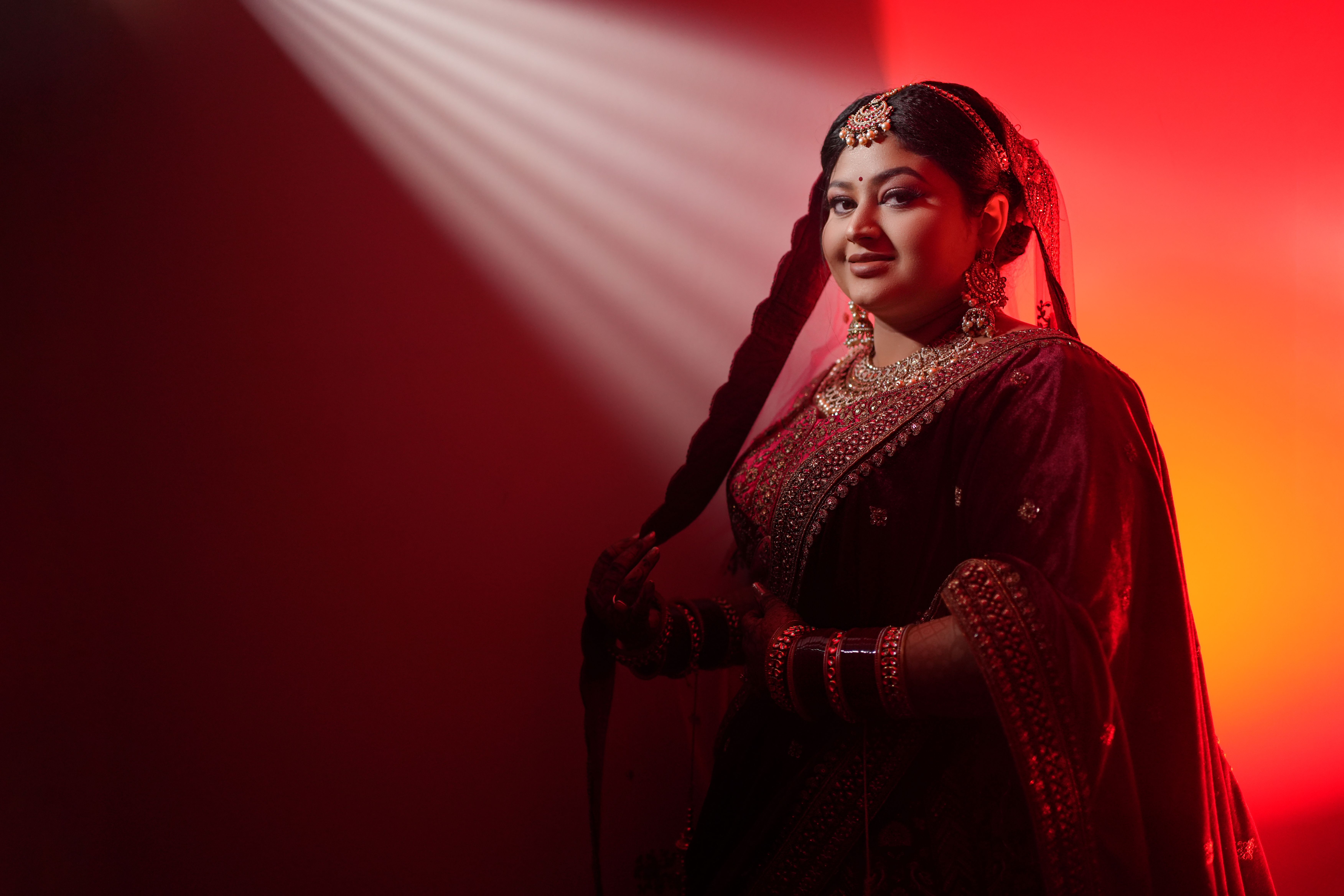Indian bride in a red gown with traditional jewelry, captured in artistic lighting.