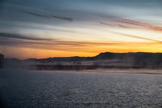 A serene sunrise over a misty lake with silhouettes of hills, creating a peaceful atmosphere.