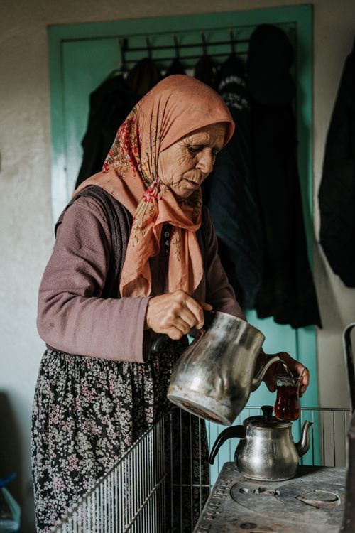 Free Elderly woman in headscarf pours tea into a kettle in a cozy kitchen setting. Stock Photo