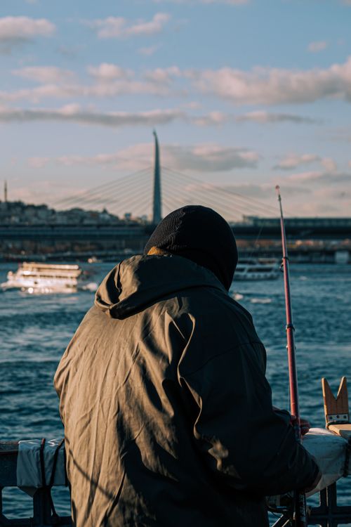 Free A person fishing from a riverside, with a bridge and boats in the background under a blue sky. Stock Photo