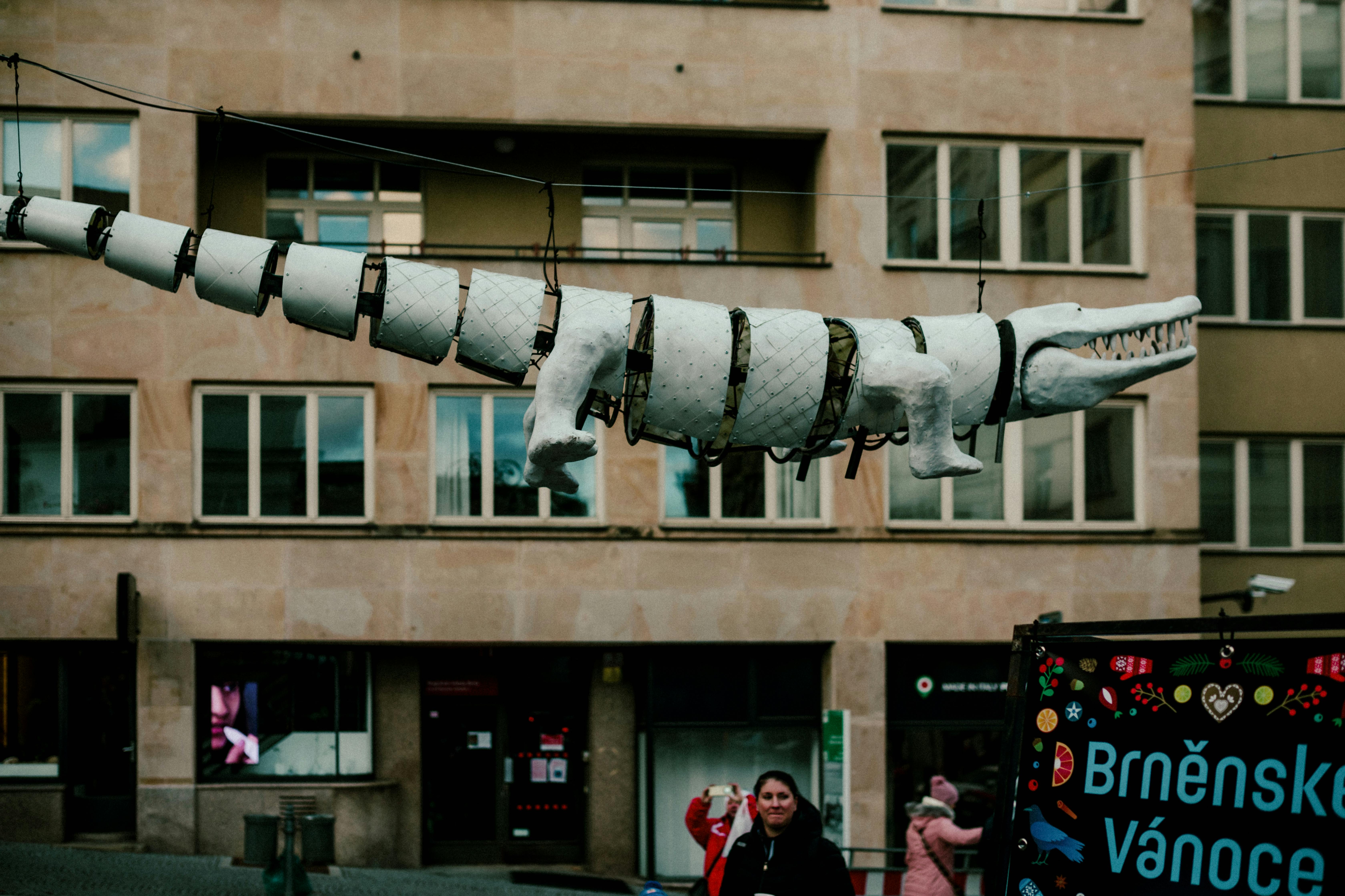 Suspended white crocodile sculpture in city with people and signage. - Brno