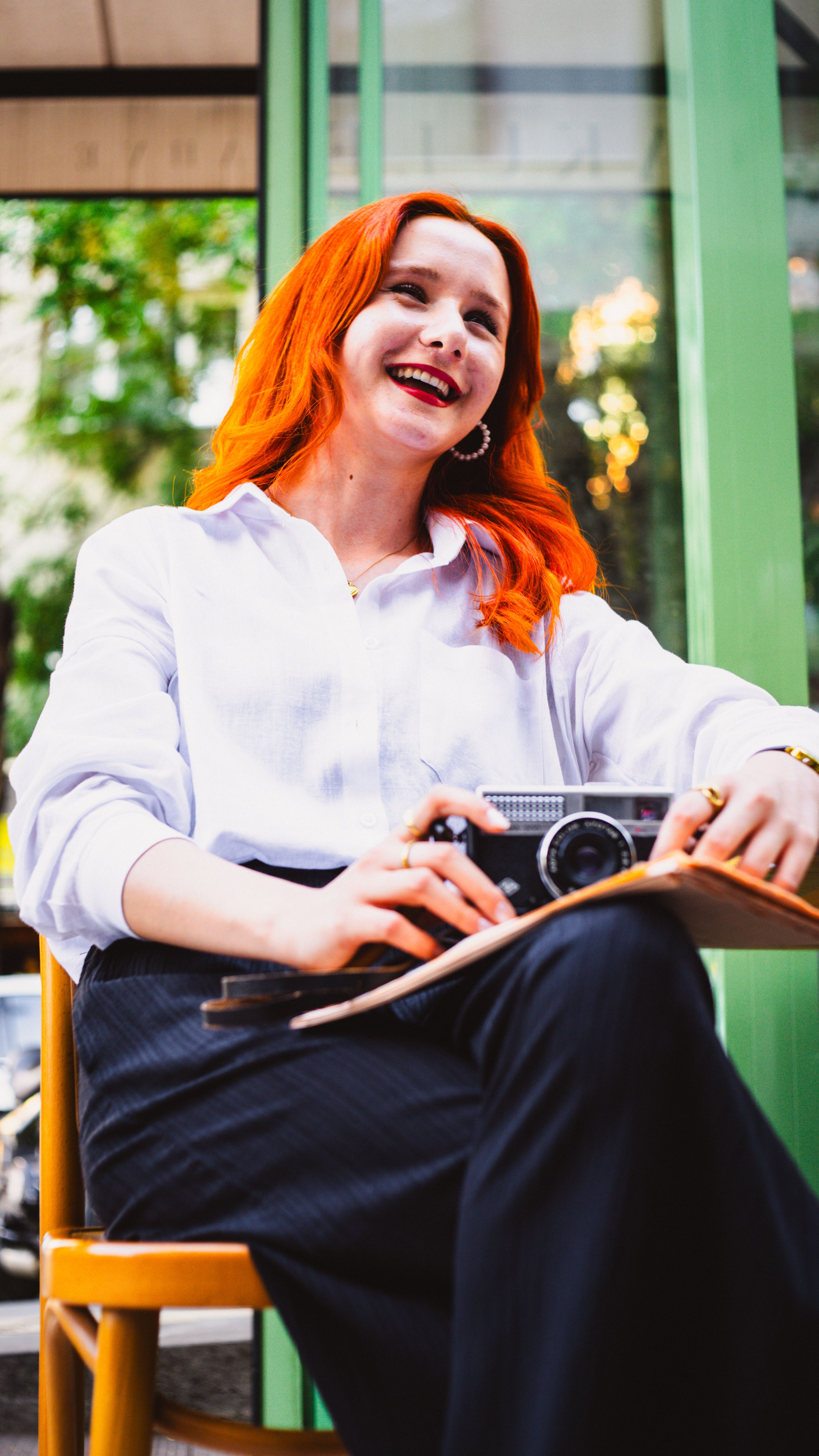 Cheerful woman with red hair holding camera outdoors, capturing joyful moments.