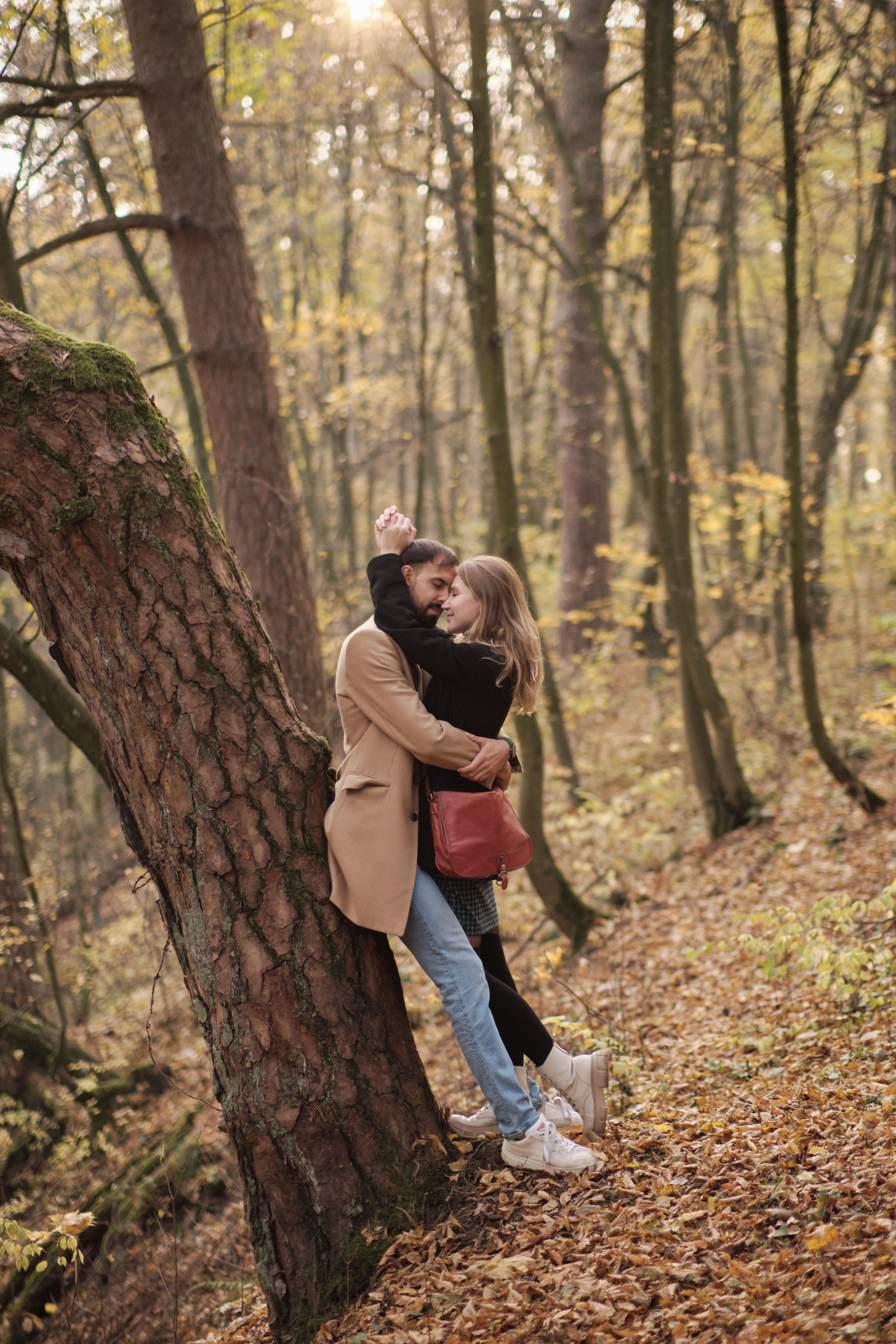 A couple lovingly embraces against a tree in a serene autumn forest setting.