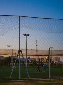 Players on a fenced soccer field practicing under stadium lights at dusk.
