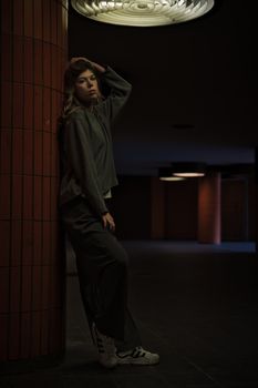 A woman leans against a tiled wall in a dimly lit urban environment, creating a moody atmosphere.
