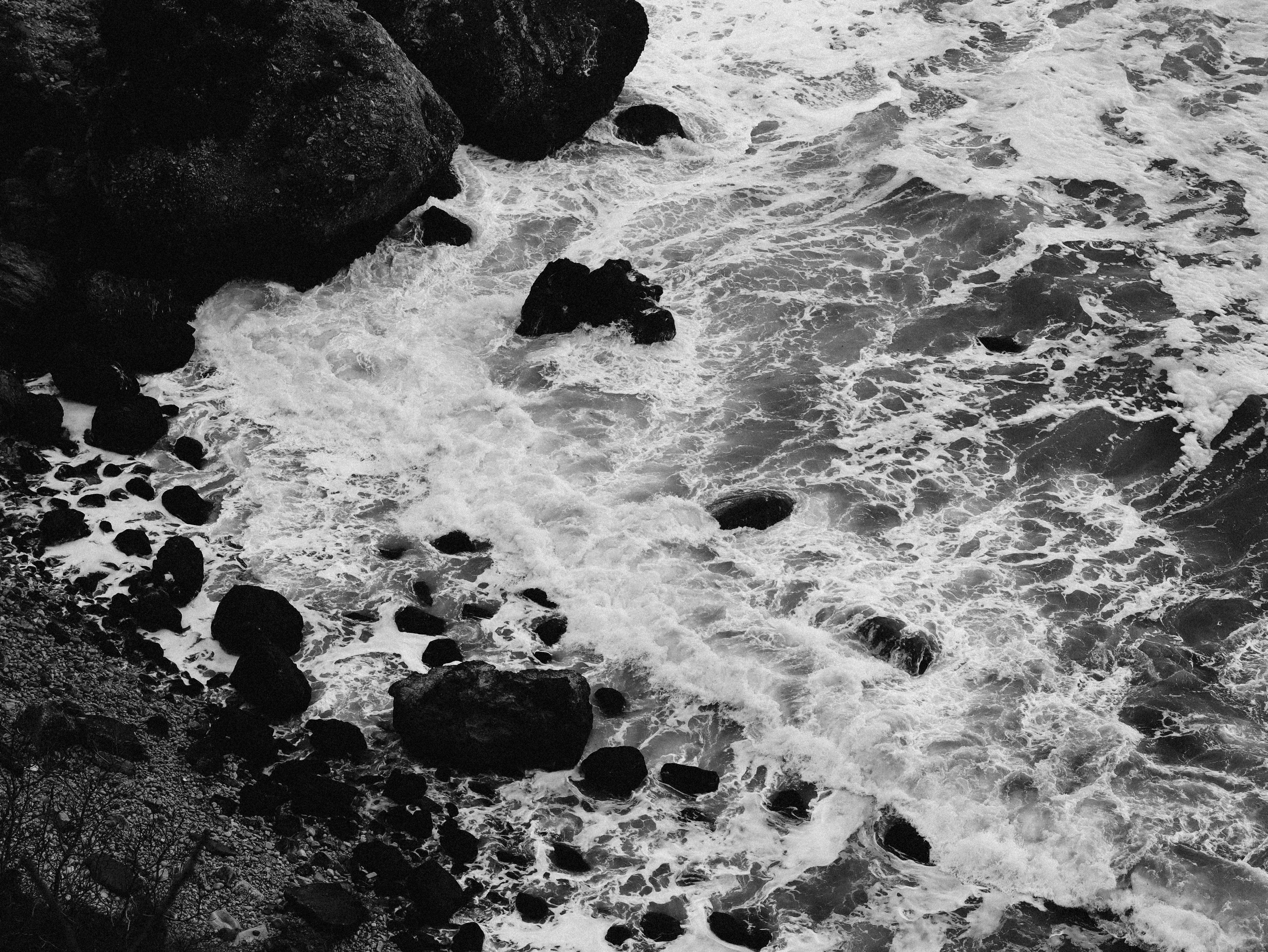 Aerial view of dark ocean waves crashing against a rugged rocky shoreline in black and white.