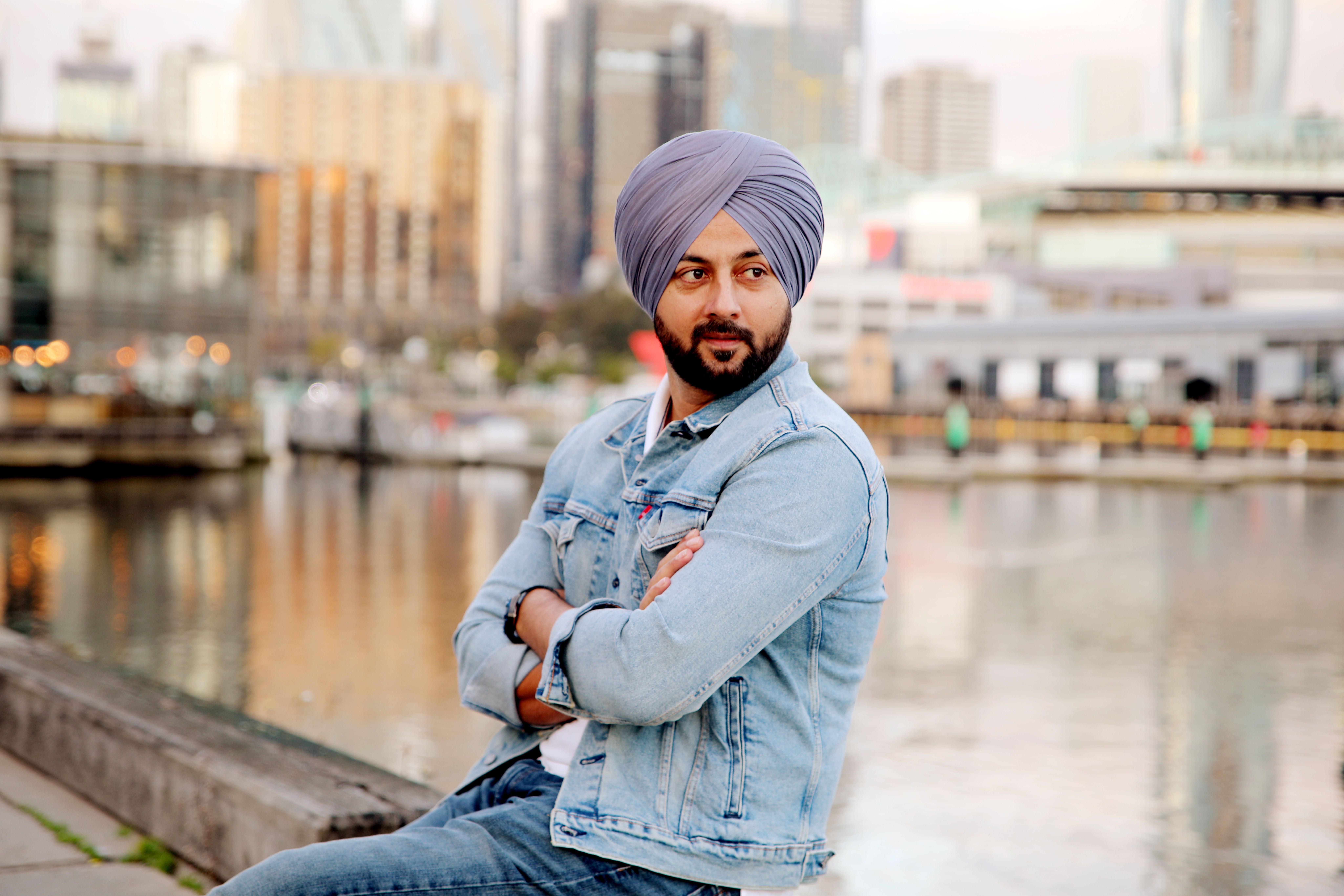 Man in denim jacket and turban relaxing by Melbourne's waterfront, urban setting.