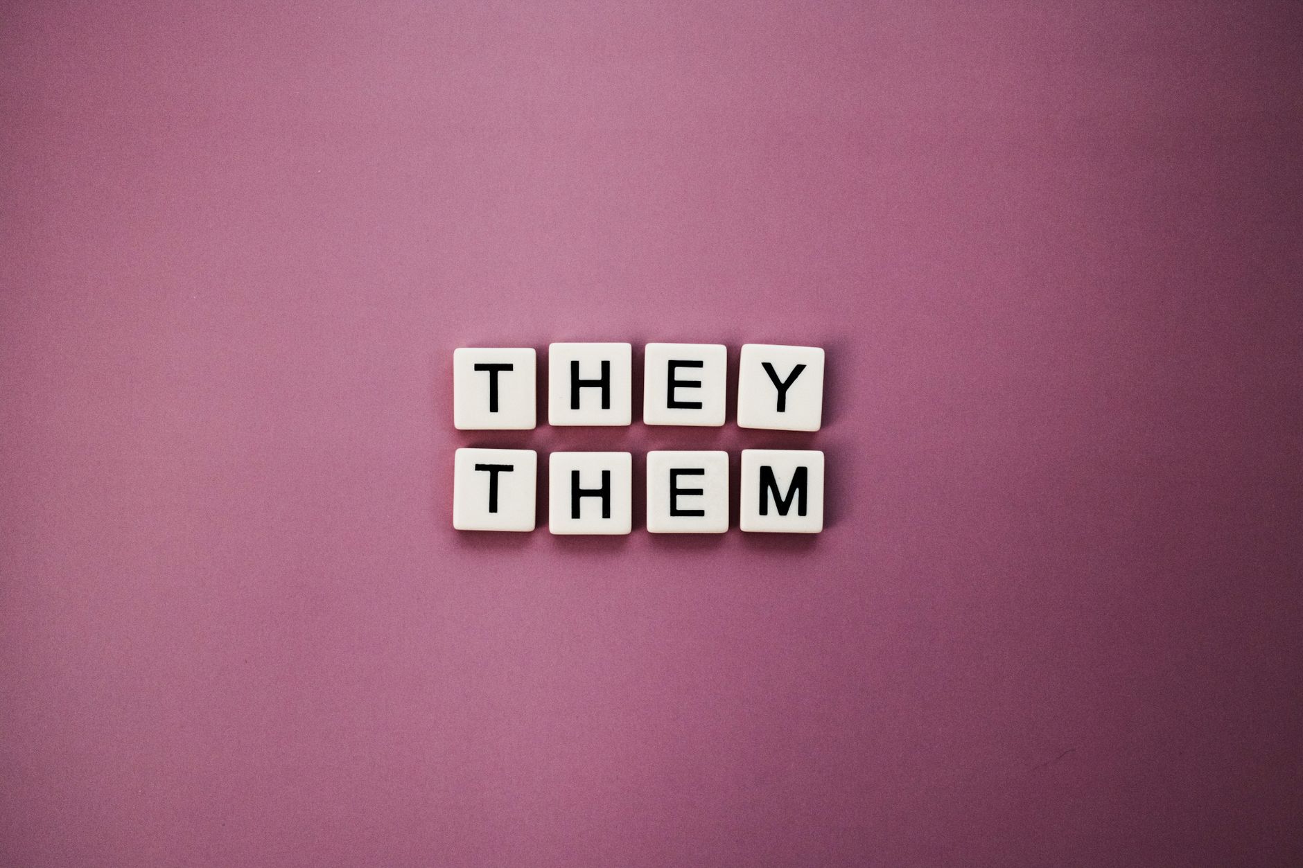 Non-binary pronouns 'they them' displayed with letter tiles on a pink backdrop.