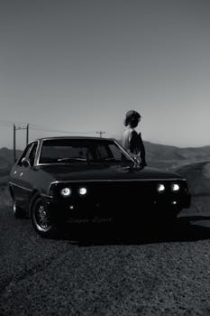 A classic car parked on a mountainous road with a silhouette person, creating a moody scene.