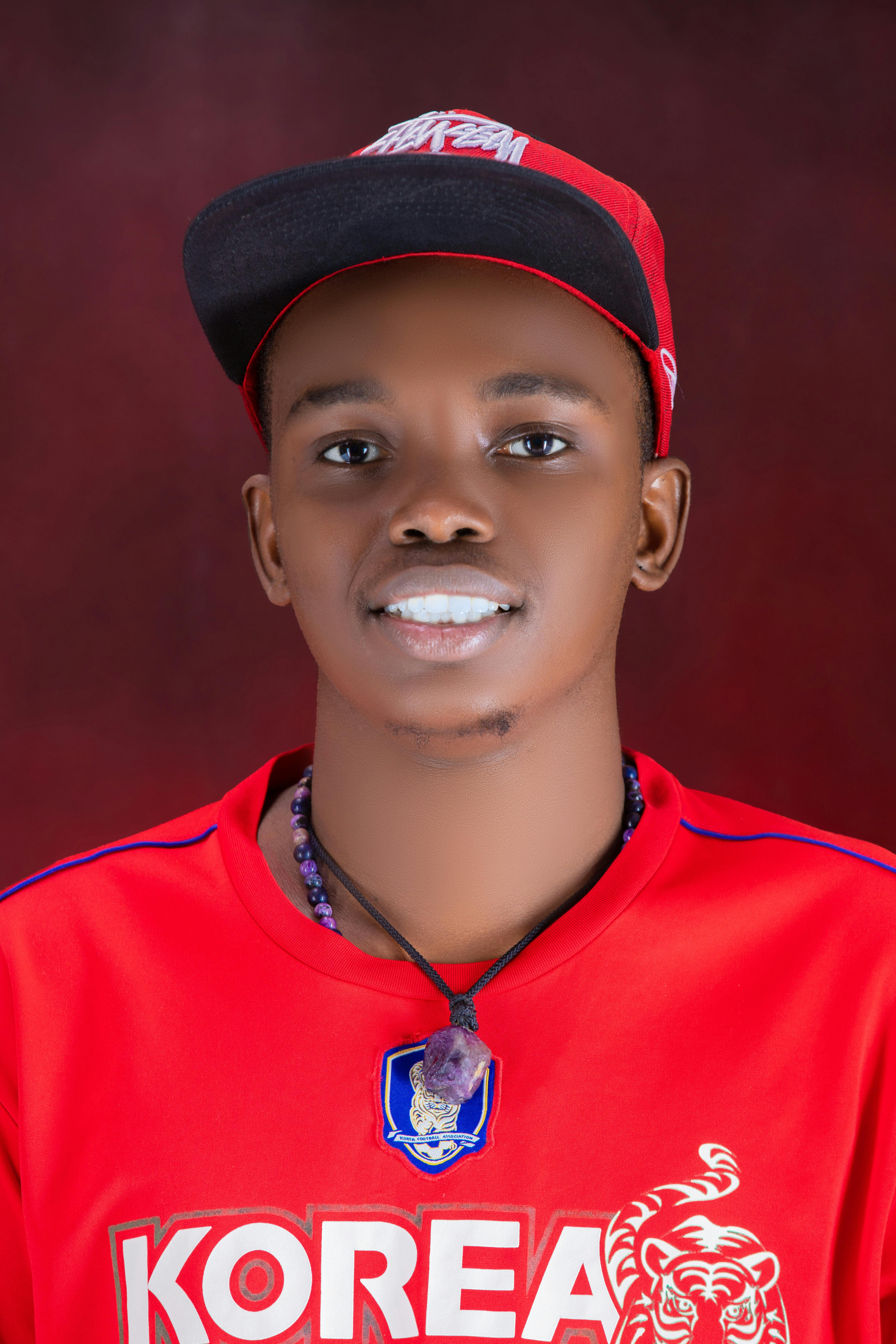 Confident young man wearing a red cap and shirt with a crystal necklace, posing against a soft background.